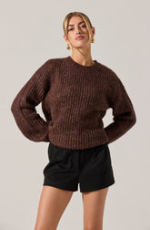 A woman is wearing a chunky, long-sleeved, brown knitted sweater with a textured pattern, and black high-waisted shorts with a loose fit. She has gold hoop earrings and styled blonde hair.